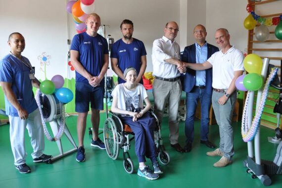 Eine Gruppe von sechs Erwachsenen steht um eine lächelnde Person im Rollstuhl in einem dekorierten Raum mit Luftballons und Trainingsgeräten. Zwei Männer schütteln sich in der Mitte die Hände, was auf ein feierliches oder unterstützendes Ereignis hindeutet.