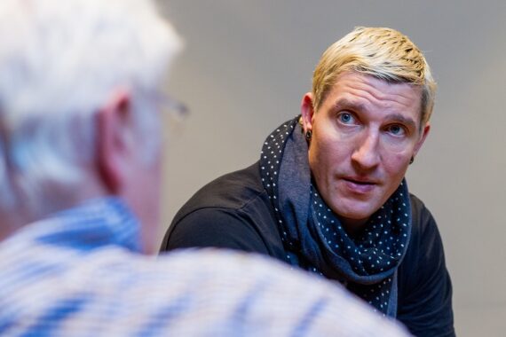 Ein blonder Mann, der einen dunklen gepunkteten Schal trägt, hört einer älteren Person mit weißem Haar und Brille, die von hinten zu sehen ist, aufmerksam zu. Der Hintergrund ist schlicht und sanft beleuchtet.