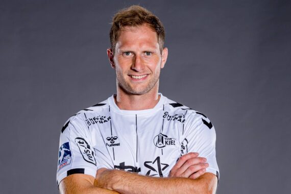 Ein männlicher Fußballspieler mit kurzen hellbraunen Haaren lächelt mit verschränkten Armen, trägt ein weißes und schwarzes Mannschaftstrikot mit Logos und steht vor einem schlichten grauen Hintergrund.