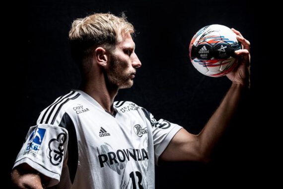 Ein männlicher Handballspieler in einem weißen Trikot hält mit einer Hand einen Handball hoch und schaut ihn vor einem dunklen Hintergrund intensiv an.