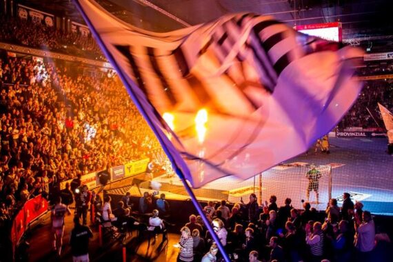 Eine große Flagge weht im Vordergrund, während eine überfüllte Sporthalle jubelt und helle Lichter und Flammen den Bereich in der Nähe des Tores erhellen. Die Spieler sind auf dem Spielfeld zu sehen, was das Ereignis noch spannender macht.