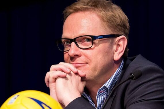 Ein Mann mit kurzen hellbraunen Haaren und Brille, der einen Anzug und ein kariertes Hemd trägt, sitzt mit unter dem Kinn verschränkten Händen da und lächelt. Im Vordergrund ist ein gelber Fußball zu sehen.