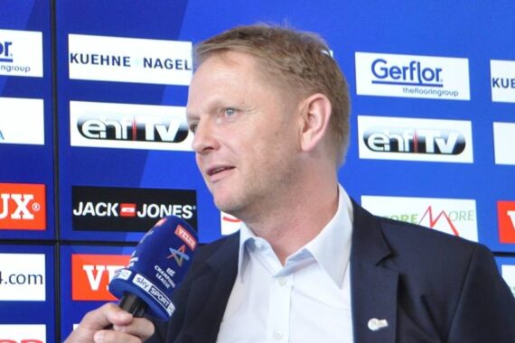 Ein Mann im Anzug wird mit einem Mikrofon vor einem Hintergrund mit verschiedenen Firmenlogos und einem blauen Farbschema interviewt.