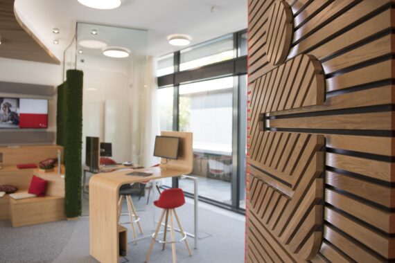 Eine moderne Bankeinrichtung mit hölzernen Akzenten, roten Stühlen, einem Computer auf einem hölzernen Schreibtisch und eingebauten Sitzgelegenheiten mit Kissen. Ein großes Holzlogo ziert die Wand im Vordergrund. Glaswände lassen natürliches Licht ins Innere.