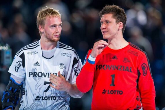 Zwei männliche Handballspieler in Mannschaftstrikots unterhalten sich auf dem Spielfeld; der eine trägt ein weißes Hemd mit der Nummer 7, der andere ein rotes Torwarthemd mit der Nummer 1. Beide wirken entspannt, das Publikum im Hintergrund ist unscharf.