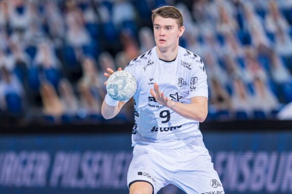 Ein männlicher Handballspieler in einem weißen Trikot mit der Nummer 9 bereitet sich darauf vor, während eines Spiels einen Ball zu fangen oder zu werfen, mit unscharfen Zuschauern auf blauen Sitzen im Hintergrund.