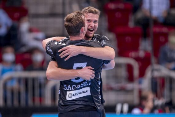 Zwei männliche Handballspieler in schwarzen Trikots umarmen sich jubelnd auf einem Hallenplatz. Der eine Spieler steht mit dem Rücken zur Kamera, während der andere freudig lächelt. Im Hintergrund sind leere rote Sitze und verschwommene Zuschauer zu sehen.