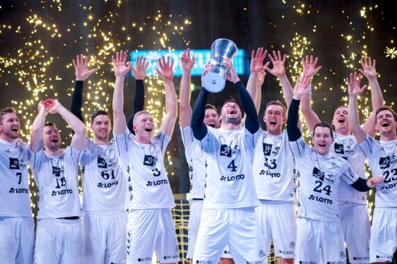 Eine Herren-Handballmannschaft in weißen Trikots feiert und hebt mit freudigen Gesichtern eine Trophäe in die Höhe, während im Hintergrund goldene Funken und Luftschlangen sprühen.
