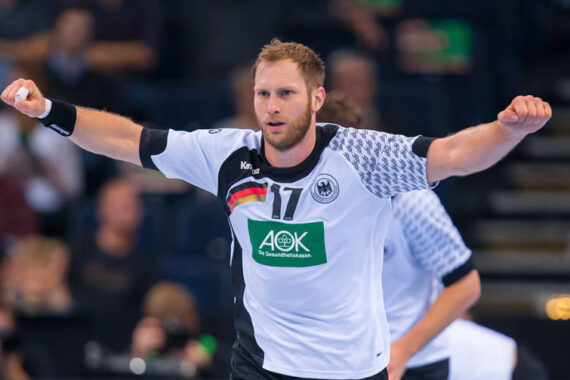 Ein männlicher Handballspieler im weiß-schwarzen Trikot der deutschen Mannschaft mit der Nummer 17 reißt auf einem Hallenplatz beide Arme jubelnd in die Höhe, während im Hintergrund unscharfe Zuschauer zu sehen sind.