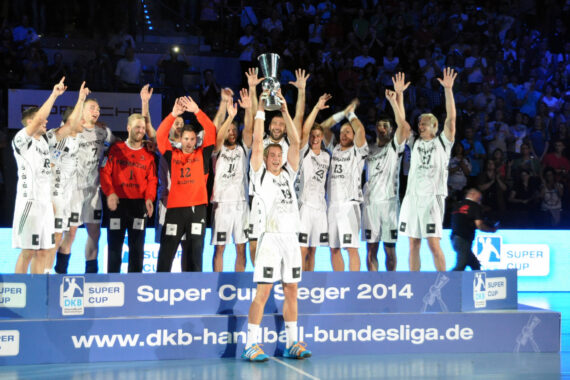 Eine Handballmannschaft der Männer feiert auf einem Podium nach dem Gewinn des DKB Handball-Bundesliga-Supercups 2014. Ein Spieler hält die Trophäe über seinen Kopf, während seine Mannschaftskameraden jubeln, während im Hintergrund eine Menschenmenge zu sehen ist.