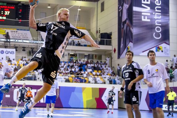 Ein Handballspieler in einem schwarzen Trikot springt, um den Ball in Richtung Tor zu werfen, während gegnerische Spieler in weißen Trikots bei einem Hallenspiel vor einer großen Menschenmenge im Hintergrund zusehen.