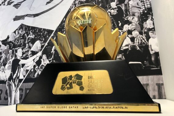 Eine goldene IHF Super Globe Qatar-Trophäe mit einem kugelförmigen Aufsatz steht auf einem schwarzen Sockel mit Logos und Aufschriften. Im Hintergrund sieht man eine schwarz-weiße Menschenmenge bei einer Veranstaltung.