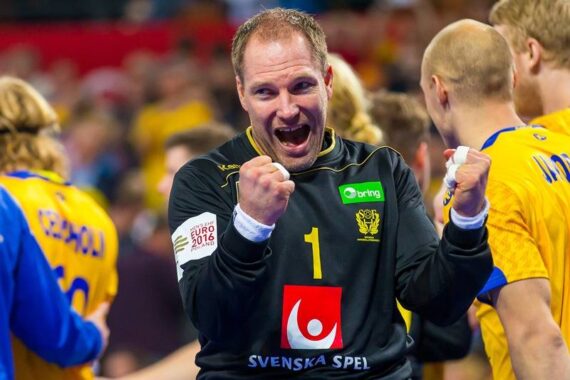 Ein männlicher Handballspieler in einem schwarzen Trikot feiert enthusiastisch mit geballten Fäusten zwischen Mannschaftskameraden in gelben Trikots auf einem Spielfeld. Die Atmosphäre ist energiegeladen und fröhlich.