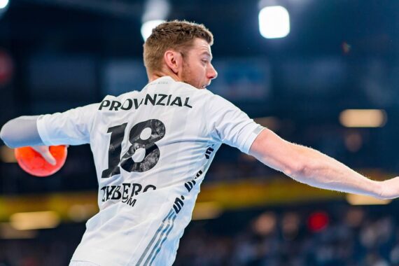 Ein männlicher Handballspieler, der ein weißes Trikot mit der Nummer 18 auf dem Rücken trägt, bereitet sich darauf vor, einen orangefarbenen Ball während eines Spiels zu werfen, mit einer unscharfen Menschenmenge und Lichtern im Hintergrund.
