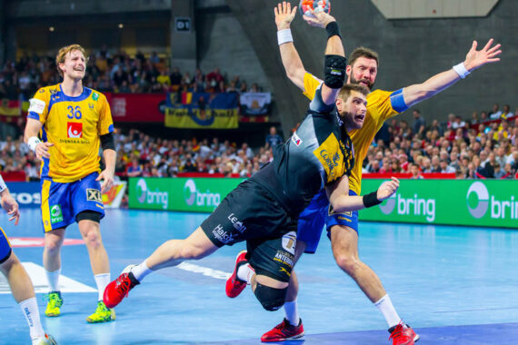 Ein Handballspieler in Schwarz befindet sich in der Luft, als er einen Schuss versucht, während ein Spieler in Gelb mit erhobenen Armen abwehrt. Das Spiel findet auf einem Hallenplatz statt, mit Zuschauern und Bannern im Hintergrund.