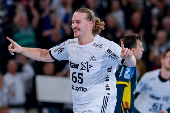 Ein männlicher Handballspieler in einem weißen Trikot mit der Nummer 65 lächelt und gestikuliert mit beiden erhobenen Zeigefingern, während er auf dem Spielfeld feiert. Im Hintergrund sind andere Spieler und eine verschwommene Menge zu sehen.