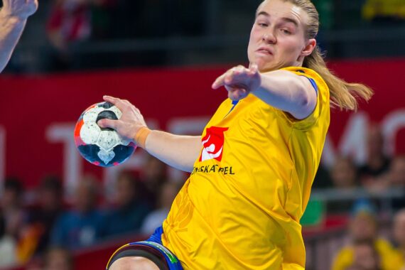 Ein Handballspieler in einem gelben Trikot springt und bereitet sich mit ausgestrecktem Arm und konzentrierter Miene auf den Wurf des Balls während eines Spiels vor.