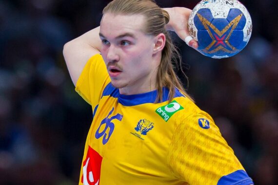 Ein männlicher Handballspieler mit langen blonden Haaren, der ein gelb-blaues Trikot trägt, bereitet sich darauf vor, einen blau-weißen Handball während eines Spiels zu werfen. Der Hintergrund ist mit Zuschauern unscharf.
