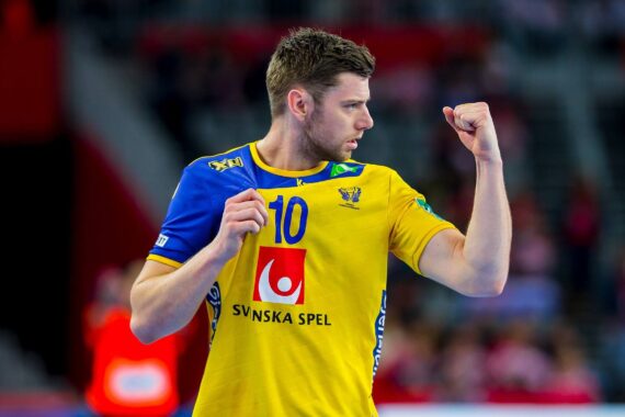 Ein männlicher Handballspieler in einem gelb-blauen Trikot mit der Nummer 10 hebt jubelnd die Faust auf einem Spielfeld, im Hintergrund sind unscharfe Zuschauer zu sehen.