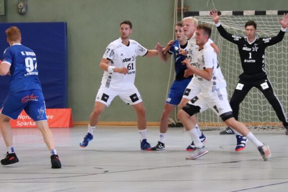 Ein Handballspieler in Blau bereitet sich auf den Wurf vor, während vier Spieler in Weiß den Ball verteidigen und ein Torwart in Schwarz wachsam vor dem Tor steht. Die Szene findet in einer Halle auf einem Sportplatz statt.