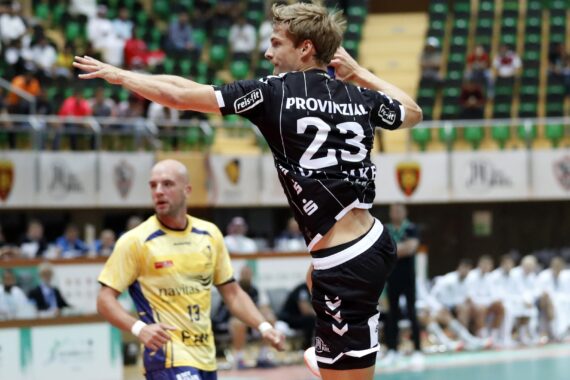 Ein Handballspieler im schwarzen Trikot mit der Nummer 23 springt, um den Ball während eines Spiels zu werfen, während ein Gegner im gelben Trikot zusieht. Das Spiel findet in einer Halle mit Zuschauern im Hintergrund statt.