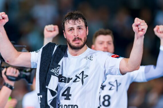 Ein männlicher Handballspieler in einem weißen Trikot hebt jubelnd beide Fäuste und hat ein Handtuch über die Schulter geworfen. Im Hintergrund sind ein weiterer Spieler und ein Kameramann zu sehen. Die Szene deutet auf einen Sieg oder ein Tor hin.