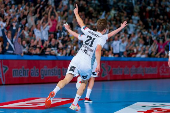 Ein Handballspieler mit der Trikotnummer 21 feiert ein Tor mit erhobenem Arm, während er auf dem Spielfeld läuft, während im Hintergrund eine begeisterte Menge jubelt.