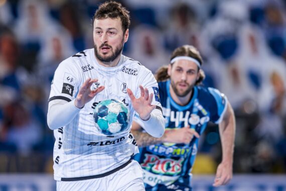 Ein männlicher Handballspieler in einer weißen Uniform bereitet sich darauf vor, einen blau-grünen Ball zu fangen, während ein Konkurrent in einer blauen Uniform ihn während eines Spiels verfolgt. Im Hintergrund ist ein unscharfes Publikum zu sehen.