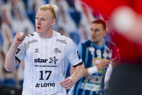 Ein Handballspieler in einem weißen Trikot mit der Nummer 17 jubelt mit geballter Faust. Ein anderer Spieler in einem blau gestreiften Trikot ist im Hintergrund auf einem Hallenplatz zu sehen.