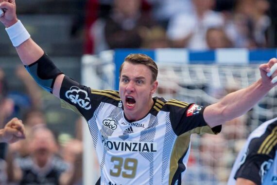 Ein männlicher Handballspieler in einem weiß-schwarzen Trikot mit der Nummer 39 jubelt mit erhobenen Armen und entschlossenem Gesichtsausdruck während eines Spiels. Der Hintergrund zeigt eine unscharfe Menge und ein Tornetz.