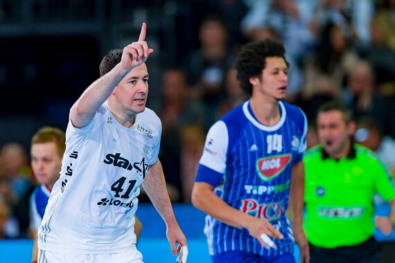 Ein Handballspieler in einem weißen Trikot hebt den Finger, während ein anderer Spieler in einem blauen Trikot und ein Schiedsrichter in Grün während eines Spiels in der Nähe des Spielfelds stehen, mit einer verschwommenen Menschenmenge im Hintergrund.