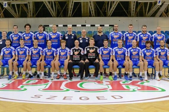 Eine Herren-Handballmannschaft in blauen Trikots posiert für ein Gruppenfoto in einer Turnhalle. Zwei Trainer und zwei Torhüter in Schwarz sitzen vorne. Ein großes Logo von Pick Szeged ist auf dem Boden zu sehen.
