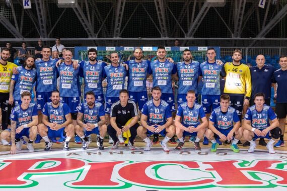 Eine Herren-Handballmannschaft posiert für ein Gruppenfoto auf einem Hallenplatz. Sie trägt blaue und gelbe Trikots, und die Trainer stehen an den Seiten. Das Spielfeld hat ein großes PICK-Logo an der Vorderseite.