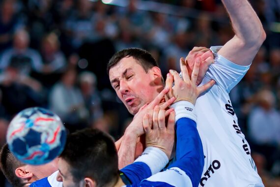 Eine Nahaufnahme von intensivem Handballgeschehen, mit Spielern in weißen und blauen Trikots, die um den Ball kämpfen, mit Händen und Gesichtern, die Entschlossenheit und Konzentration inmitten einer verschwommenen Menge zeigen.