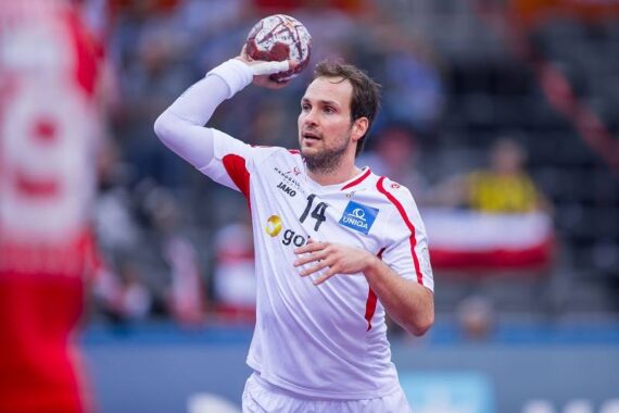 Ein männlicher Handballspieler in einem weiß-roten Trikot mit der Nummer 14 bereitet sich darauf vor, den Ball während eines Spiels zu werfen. Der Hintergrund ist unscharf, ein anderer Spieler in Rot ist teilweise sichtbar.