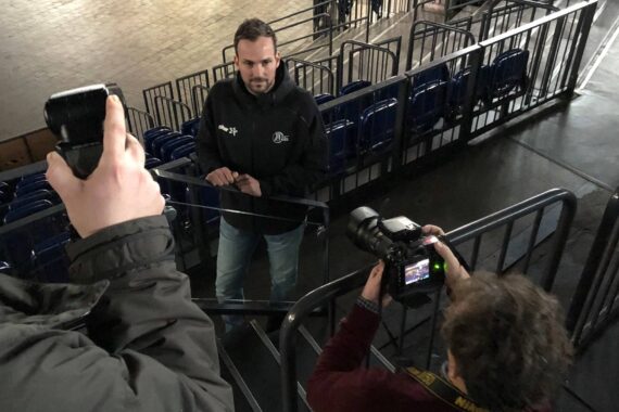 Ein Mann steht auf einer Treppe in einer Arena und sieht sich zwei Fotografen gegenüber - einer hält eine Kamera mit Blitzlicht, der andere fotografiert ihn von unten. Im Hintergrund sind blaue Sitze und Metallgeländer zu sehen.
