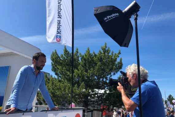 Ein Mann in einem blauen Hemd posiert im Freien, während ein anderer Mann sein Foto mit einer professionellen Kamera und Beleuchtungsausrüstung aufnimmt. Im Hintergrund sind Bäume, eine Flagge und ein klarer blauer Himmel zu sehen.