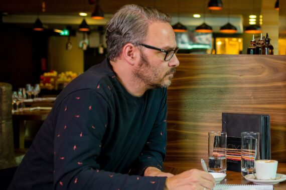Ein Mann mit Brille und grauem Haar sitzt an einem Tisch in einem Restaurant und schreibt in ein Notizbuch. Zwei Gläser Wasser und eine Speisekarte stehen auf dem Tisch. Warme Beleuchtung und unscharfer Hintergrund schaffen eine gemütliche Atmosphäre.
