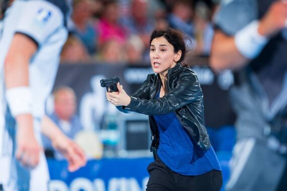 Eine Frau in einer schwarzen Lederjacke richtet mit konzentrierter Miene eine Handfeuerwaffe nach vorne. Sie ist umgeben von Menschen in Sportkleidung, und die Szene wirkt angespannt und dramatisch.