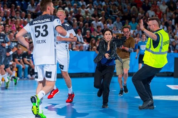 Eine Frau rennt während eines Spiels auf ein Handballfeld und wird von einem Sicherheitsbeamten aufgehalten, während Spieler und ein Kameramann zusehen. Das Spielfeld ist mit Zuschauern gefüllt.
