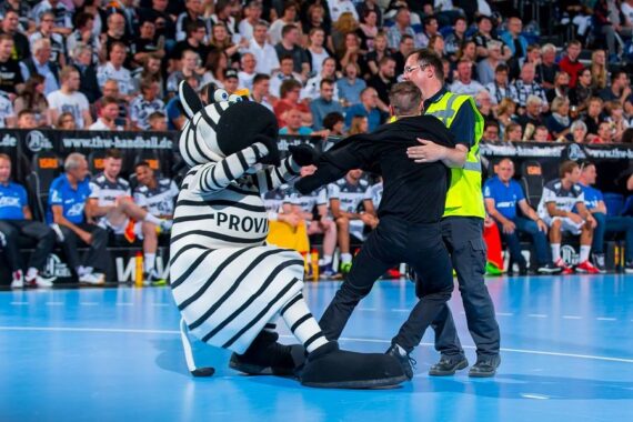 Eine Person in einem Zebra-Maskottchenkostüm sitzt auf dem Boden, während ein Sicherheitsbeamter versucht, eine andere Person während einer Sportveranstaltung wegzuziehen, während im Hintergrund eine Menschenmenge zusieht.