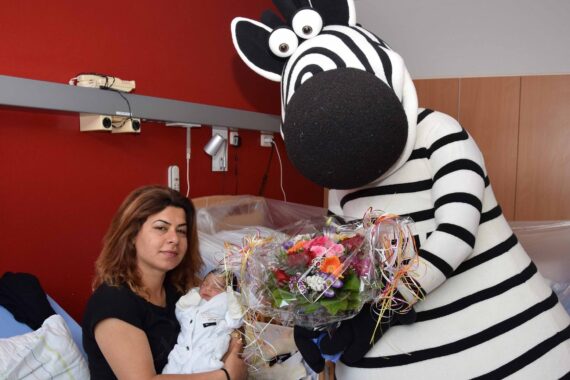 Eine Frau mit einem Baby im Arm erhält in einem Krankenhauszimmer einen bunten Blumenstrauß von einer Person in einem großen Zebrakostüm. Die Umgebung wirkt fröhlich und feierlich.