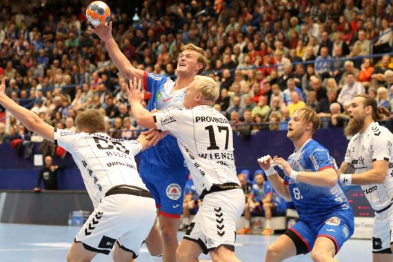 Ein Handballspieler in einem blauen Trikot springt mit dem Ball, während drei gegnerische Spieler in Weiß versuchen, ihn zu blockieren, während im Hintergrund eine volle Arena mit Zuschauern zu sehen ist.