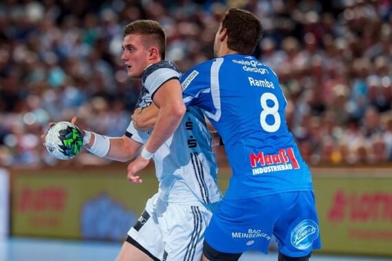 Zwei männliche Handballspieler in Aktion während eines Spiels; einer in einer weiß-grauen Uniform hält den Ball, während er von einem anderen Spieler in einer blauen Uniform mit der Nummer 8 eng verteidigt wird. Im Hintergrund ist eine Menschenmenge zu sehen.