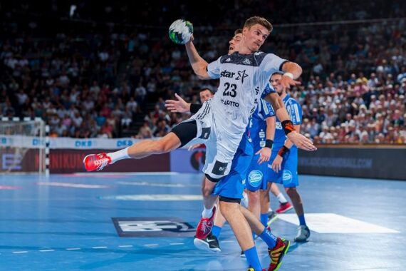 Ein Handballspieler in einem weißen Trikot springt und bereitet sich auf den Wurf vor, während er von Spielern in blauen Trikots auf einem Hallenplatz verteidigt wird, während im Hintergrund eine Menschenmenge zusieht.
