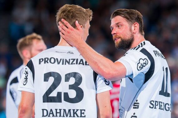 Ein Handballspieler mit der Trikotnummer 10 hält während eines Spiels sanft den Kopf eines Mannschaftskameraden mit der Trikotnummer 23, beide in weißen Trikots, um ihn zu ermutigen und zu unterstützen.