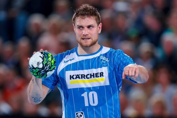 Ein männlicher Handballspieler in einem blau gestreiften Trikot mit der Nummer 10 hält während eines Spiels einen Handball in der einen Hand und gestikuliert mit der anderen, während im Hintergrund eine verschwommene Menschenmenge zu sehen ist.