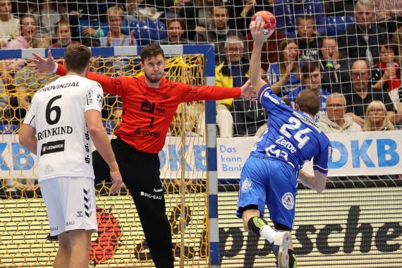 Ein Handballspieler in Blau springt und schießt den Ball in Richtung Tor, während ein Torhüter in Rot und ein Verteidiger in Weiß versuchen, ihn zu blockieren; eine Menschenmenge schaut von der Tribüne hinter dem Tornetz zu.