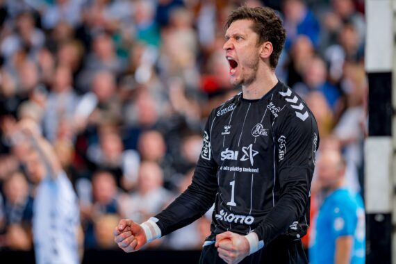 Ein Handballtorhüter in einem schwarzen Trikot ballt die Fäuste und schreit vor Begeisterung, während er ein Spiel feiert. Im Hintergrund sind eine unscharfe Menge und Spieler zu sehen.