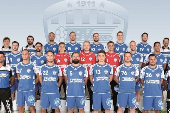 Eine Handballmannschaft der Männer posiert für ein Gruppenfoto in blau-weißen Trikots, während Trainer und Betreuer an den Seiten stehen. Das Mannschaftslogo und die Jahreszahl 1911 sind im Hintergrund schwach zu erkennen.